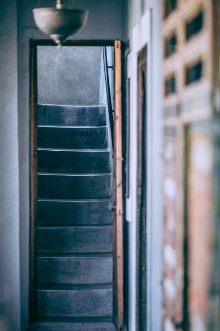 A narrow staircase with dark, textured steps inside a property, seen through an open doorway framed by wooden edges, leading upwards. The staircase is enclosed within a tight space, with a rounded, grey planter visible at the top of the image suggesting a small landing or hallway area. To the right of the staircase, a partially visible wall or partition is composed of wooden slats, providing a semi-open appearance. The environment indicates an interior space prepared for a home relocation or moving process, with the staircase likely to be a challenging feature for removals. The lighting appears natural, filtering through the surrounding areas, and the scene captures the initial stages of furniture transport or packing before removal van services, such as those provided by Removal Van Hackney, are carried out. The image emphasizes space constraints often encountered during furniture transport in urban settings like Dalston E8, highlighting the importance of professional removals to navigate narrow staircases.