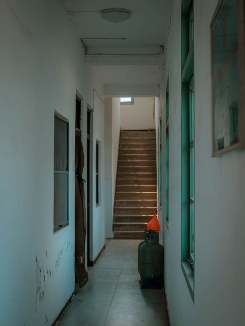 A narrow indoor corridor with white walls shows signs of wear and minor damage. On the left side, there are three small, rectangular windows with glass panes, and a brown fabric curtain hanging beside one of them. On the right side, there are large windows with green-tinted glass and wooden frames, allowing natural light to illuminate the space. The floor is tiled with light grey tiles. Near the end of the corridor, close to a wooden staircase with visible steps leading upwards, a black portable gas heater with an orange safety cone placed on top stands next to the wall. Adjacent to the staircase, a brown leather bag hangs on a wall hook. The ceiling appears slightly stained and damaged, consistent with a property undergoing a home relocation or packing process, which Removal Van Hackney may support with furniture transport and loading services for house removals.