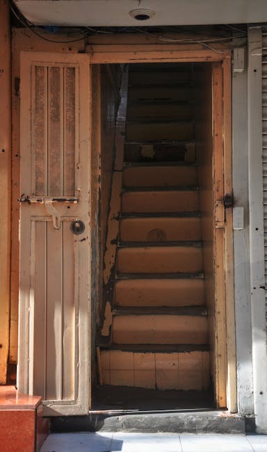 A narrow staircase with dark, textured steps inside a property, seen through an open doorway framed by wooden edges, leading upwards. The staircase is enclosed within a tight space, with a rounded, grey planter visible at the top of the image suggesting a small landing or hallway area. To the right of the staircase, a partially visible wall or partition is composed of wooden slats, providing a semi-open appearance. The environment indicates an interior space prepared for a home relocation or moving process, with the staircase likely to be a challenging feature for removals. The lighting appears natural, filtering through the surrounding areas, and the scene captures the initial stages of furniture transport or packing before removal van services, such as those provided by Removal Van Hackney, are carried out. The image emphasizes space constraints often encountered during furniture transport in urban settings like Dalston E8, highlighting the importance of professional removals to navigate narrow staircases.