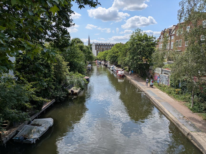 A photograph taken during daytime shows a narrow waterway in Hackney lined with green trees on both sides, with some overhanging branches casting shadows on the water. On the left side, there are boats moored along the bank, one covered with a tarp. The right side features a paved footpath with a few pedestrians walking, adjacent to multi-story brick buildings with large windows, some decorated with graffiti. Parked cars are visible along the street near the canal. Beyond the trees and buildings, a church spire and additional residential structures can be seen in the distance under a partly cloudy sky. This scene captures a typical residential area in Hackney, highlighting urban living, outdoor space, and local transportation, consistent with the themes of home relocation and moving logistics supported by Removal Van Hackney.