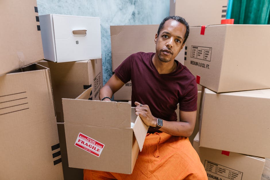 A man with dark skin, short curly hair, and wearing a maroon T-shirt and orange trousers sits among several unpacked cardboard boxes inside a property, preparing to load or organize items for a home relocation. He is holding an open cardboard box labeled 'Fragile' with red and white stickers, indicating careful handling of delicate contents. Around him, other sealed and partially opened boxes are stacked, some with black striped patterns and printed labels, suggesting organized packing and moving procedures. The room features light-colored walls with visible patches of blue and teal, and the lighting appears natural, illuminating the scene evenly. This setup reflects the logistics of furniture transport and packing involved in professional removals services. The image is relevant to house removals and moving logistics as provided by Removal Van Hackney, illustrating the careful handling of household items during the loading process.
