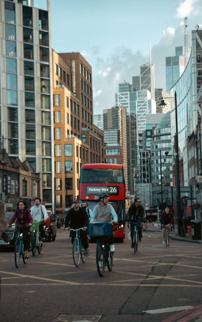A busy urban street scene in Hackney with a red double-decker bus displaying the route number 26 and destination 'Hackney Wick.' Several pedestrians and cyclists are crossing the road, some riding bicycles and others walking, all dressed in casual clothing suitable for daytime. On the left, there are a few vehicles, including a taxi, and in the background, modern glass and brick office or residential buildings rise into a partly cloudy sky. The scene depicts a typical city environment where people commute on foot and by bike, with the road marked by crossing lines and traffic signals. Although the image does not directly show house removals or moving activities, it relates to urban logistics and transport, which are part of the broader context of home relocation services offered by Removal Van Hackney, a professional removals company focused on local moving and furniture transport within Hackney.