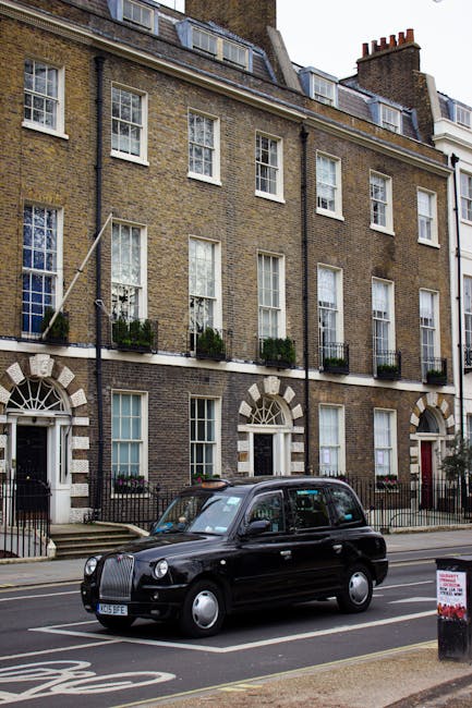 A black London taxi cab is parked on the street in front of a multi-storey residential building with classic brick facade, white framed sash windows, and small balconies with potted plants, during daylight hours. The vehicle is positioned near the curb, with the front facing left. In the background, the building features several floors with windows and chimney stacks visible on the roof. The scene suggests a typical urban environment suitable for house removals and furniture transport. The presence of the cab indicates potential involvement in home relocation or moving logistics, which Removal Van Hackney services as part of their removals offering for Hackney residents. The image captures the exterior of the building, the parked vehicle, and the street setting, emphasizing the urban context of moving services and household furniture transport.