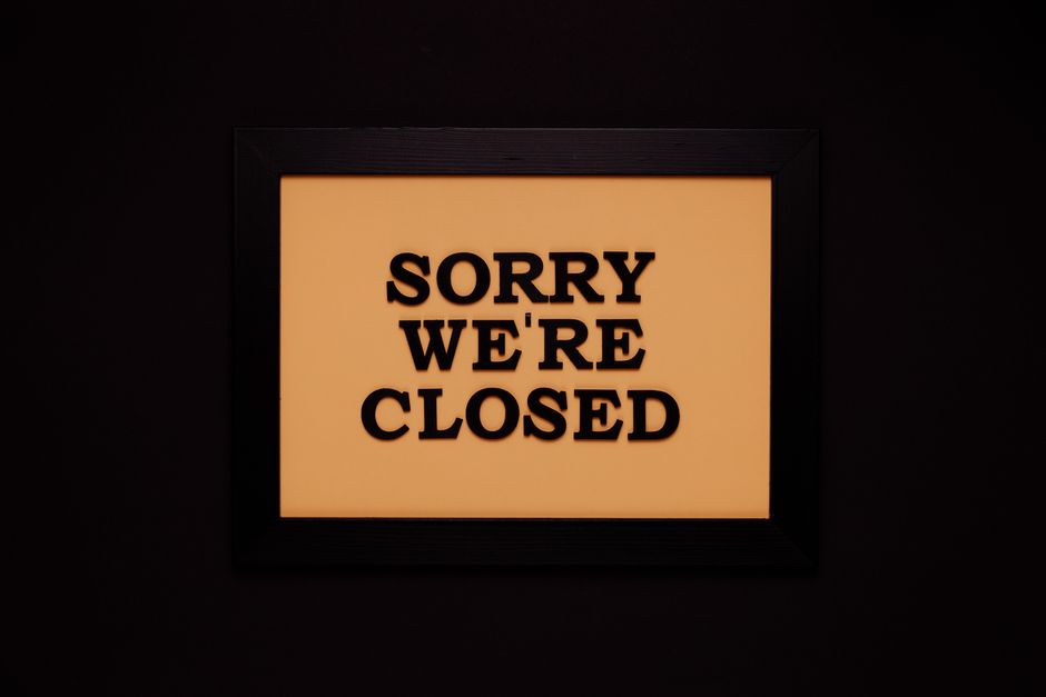 A backlit sign with an orange background and black bold uppercase letters that reads 'SORRY WE'RE CLOSED', mounted on a dark wall. The sign appears to be indoors, possibly on the exterior of a shop or